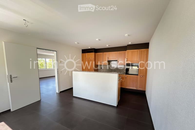 Modern kitchen with light wooden cabinets, dark tiled floor, and a white countertop. An open doorway leads to another room with large windows.