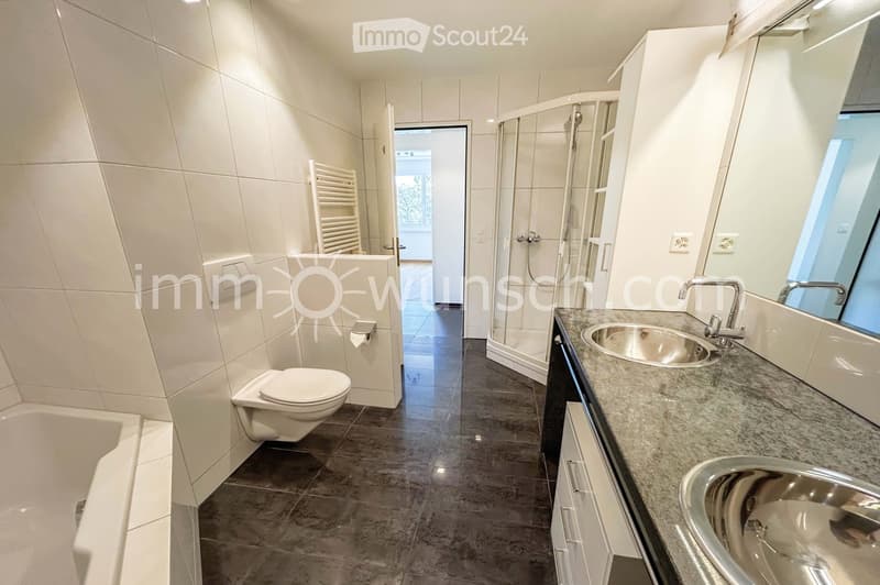 Modern bathroom with white tiles, a bathtub, toilet, double sink with chrome faucets, a large mirror, and a glass-enclosed shower. A radiator is visible on the wall.