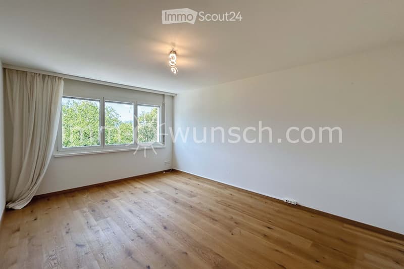 Empty room with wooden flooring, white walls, large windows with curtains, and a ceiling light fixture.
