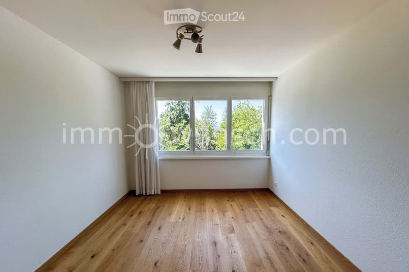 Empty room with wooden flooring, white walls, large window with curtains, and a ceiling light fixture.