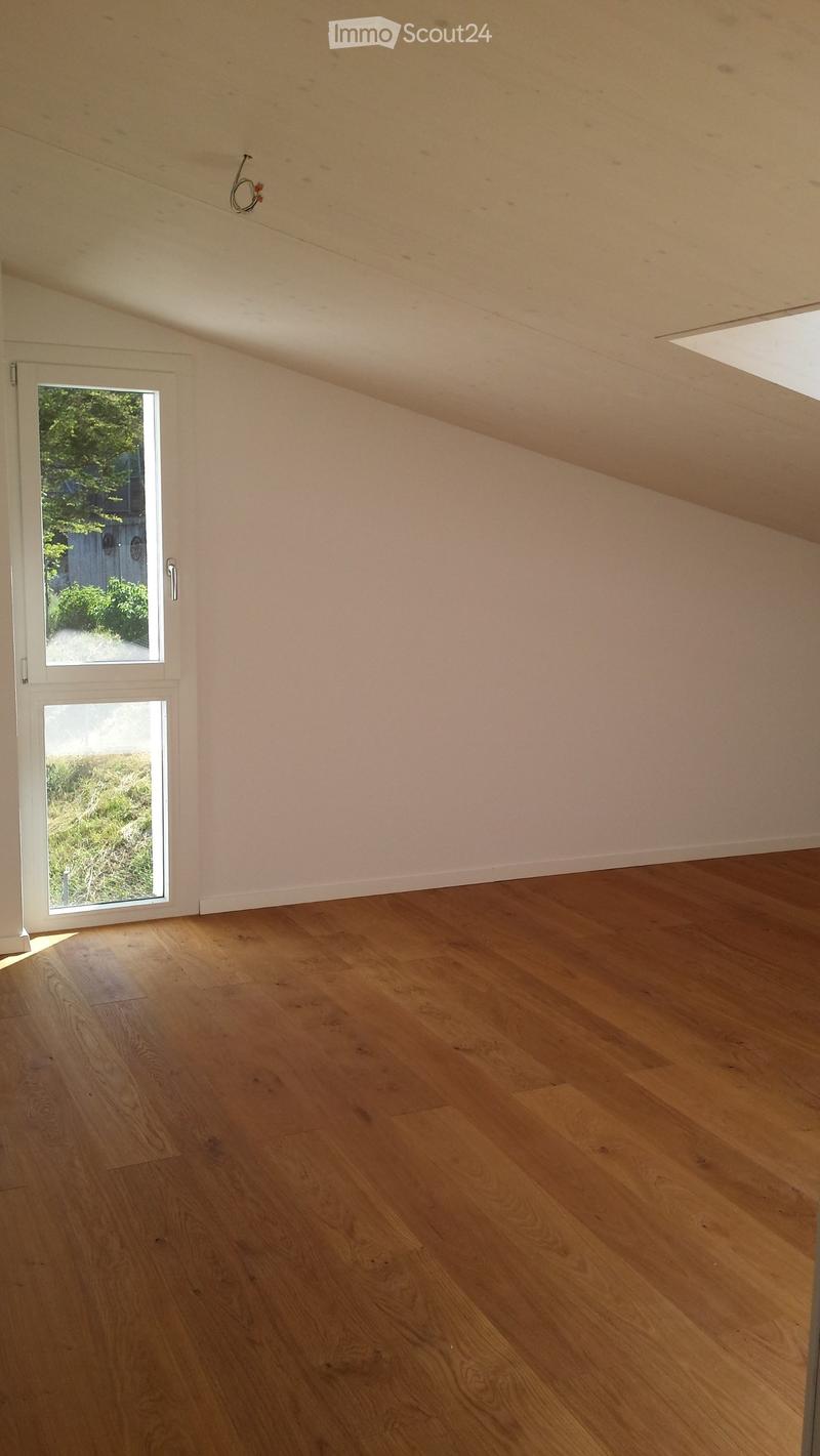 Empty room with wooden flooring, white walls, and a large window with a view of greenery outside.