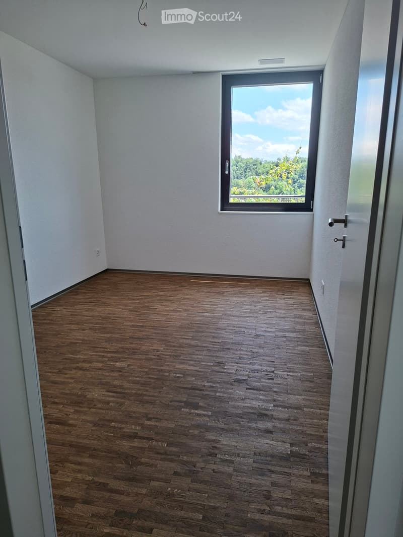 Empty room with wooden flooring, white walls, and a large window offering a view of greenery outside.