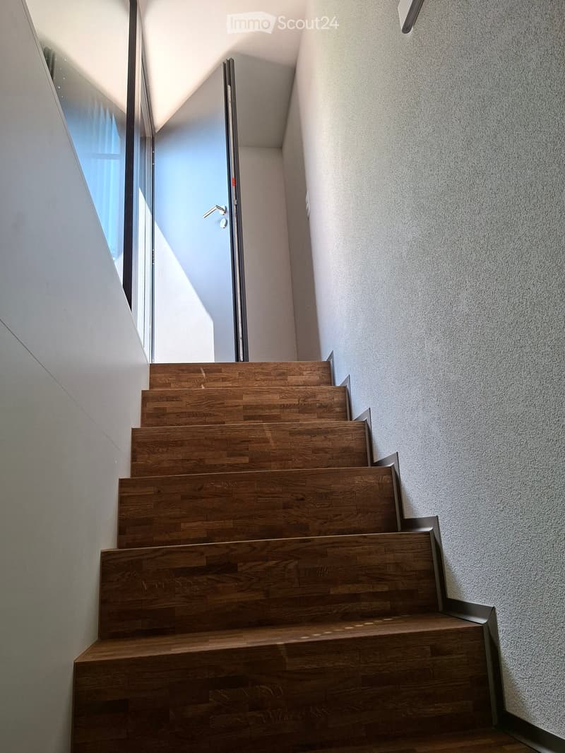 Modern staircase with wooden steps and metal railings, leading to a bright, open area with large windows and a sleek door.