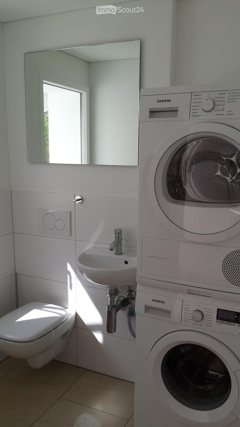 Modern bathroom with a white sink, toilet, washing machine, and a large mirror above the sink. The room features light-colored tiles and a window allowing natural light.
