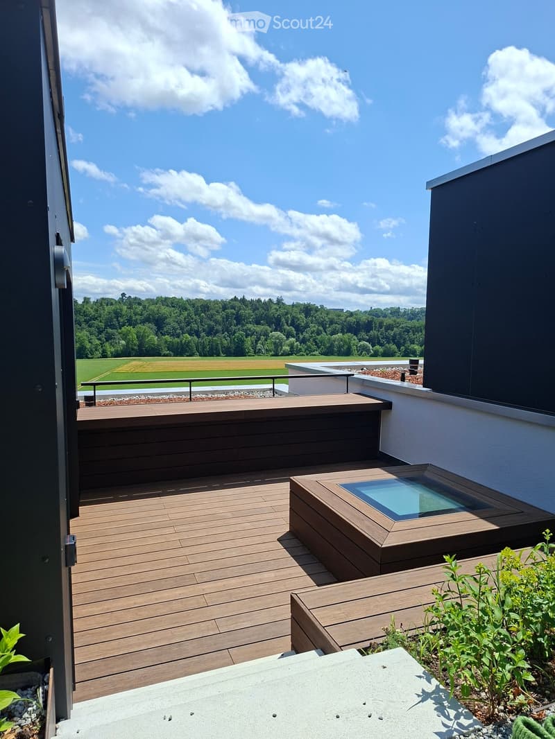 Wooden deck terrace with a glass-enclosed seating area, overlooking a green landscape with trees and fields.