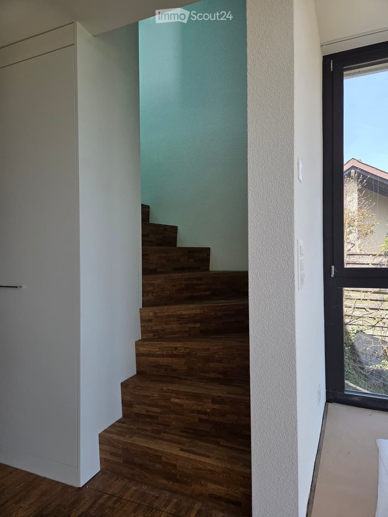 A modern hallway with wooden flooring, a staircase leading upwards, and a large window offering a view of the outdoor area.