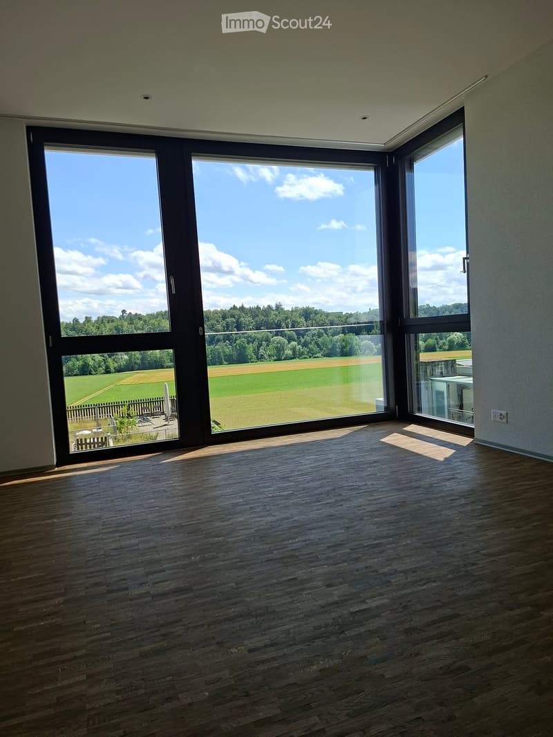 Empty room with wooden flooring and large windows offering a view of a green field and trees.