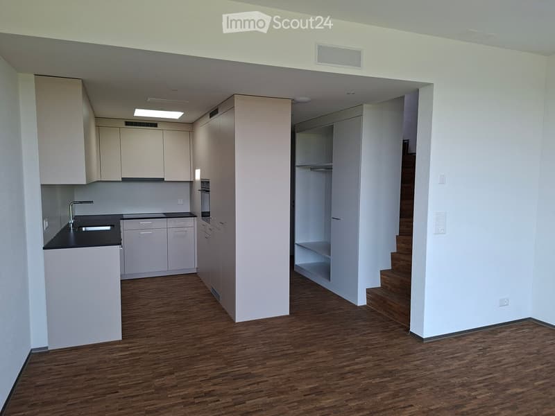 Empty room with wooden flooring, white walls, and a small kitchen area with cabinets and a sink. A staircase leads to another level.