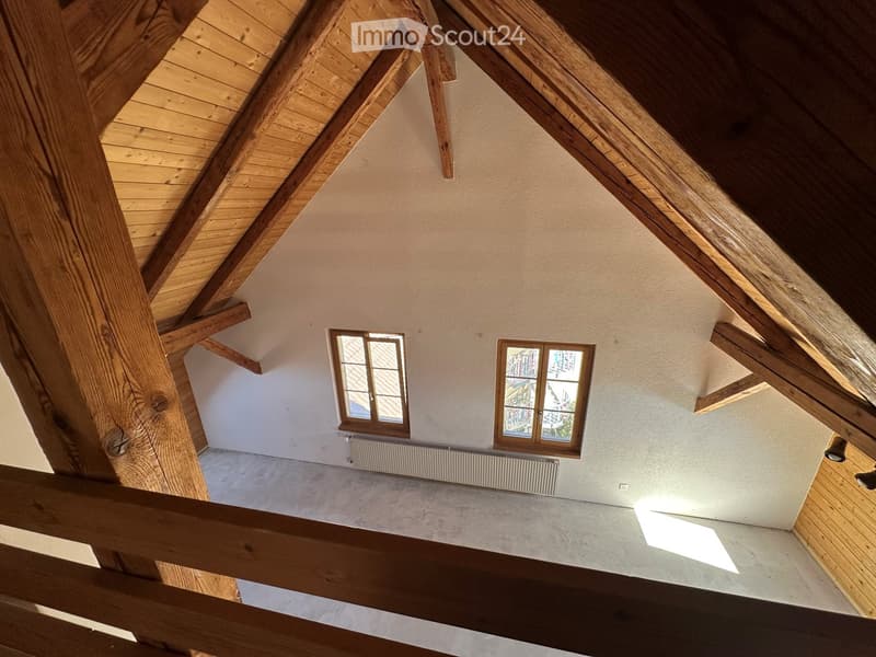 Empty attic space with wooden beams, exposed wooden ceiling, two windows, and a radiator. No furniture or equipment present.