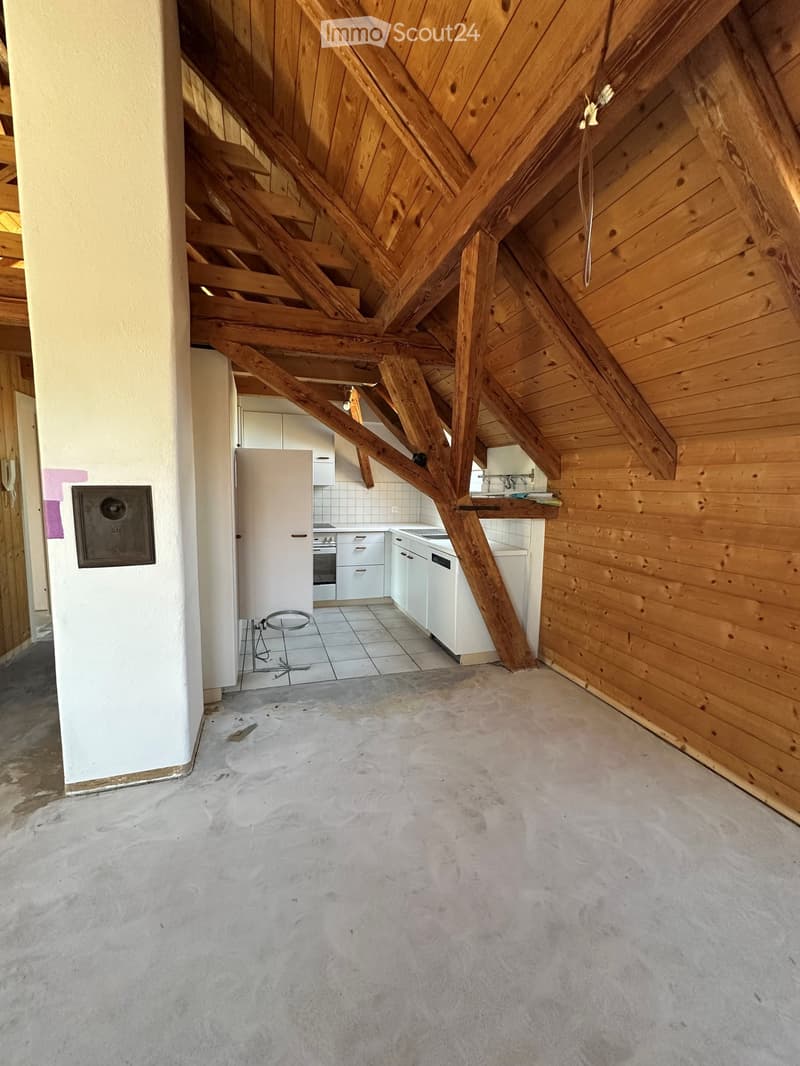 An attic space with exposed wooden beams and a concrete floor, featuring a small kitchen area with white cabinets and appliances in the background.