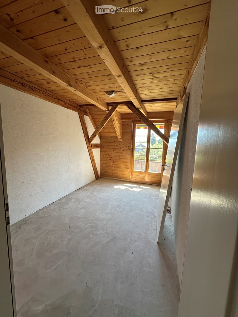 Empty room with exposed wooden beams and a concrete floor, featuring large windows and a door leading outside.