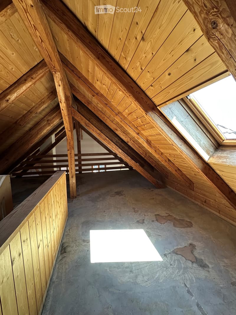 An attic space with exposed wooden beams and rafters, unfinished concrete floor, and a small skylight window allowing natural light to enter.