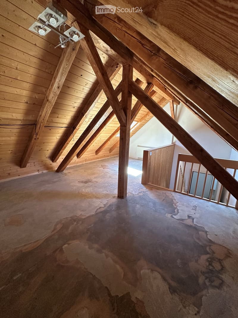 An attic space with exposed wooden beams and a slanted wooden ceiling. The floor is unfinished concrete, and there is a wooden door leading to another area.