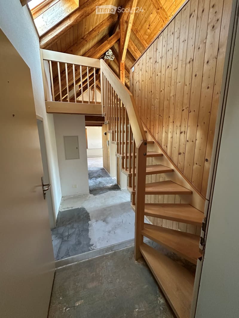 Wooden staircase with a handrail leads to an attic space with exposed wooden beams and skylight. The hallway has a concrete floor and white walls.