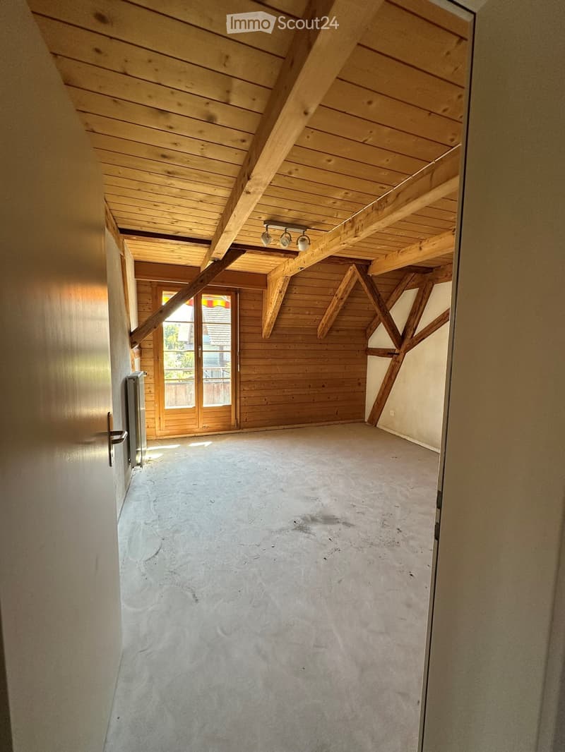 Empty room with exposed wooden beams and a concrete floor, featuring a large window and a door leading outside.
