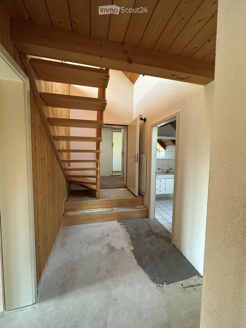 Wooden staircase with open wooden railings leads to a hallway with light-colored walls and a concrete floor. A bathroom with white cabinets and a sink is visible to the right.