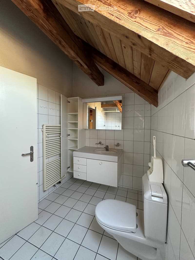 Modern bathroom with white tiles, a toilet, a sink with a cabinet, a mirror, and a radiator. The ceiling has exposed wooden beams.