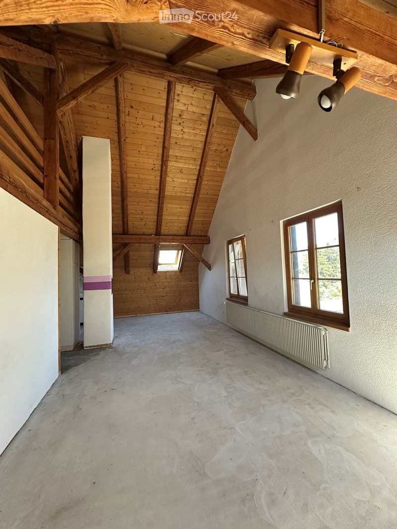 Empty room with exposed wooden beams, concrete floor, and large windows letting in natural light. No furniture or equipment present.