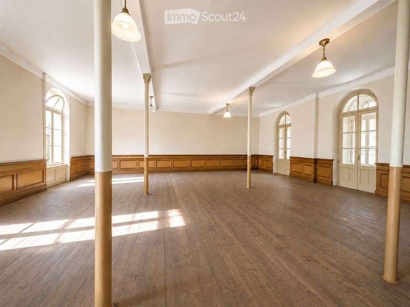 Empty room with wooden flooring, white walls, large arched windows, and central columns. No furniture or equipment present.