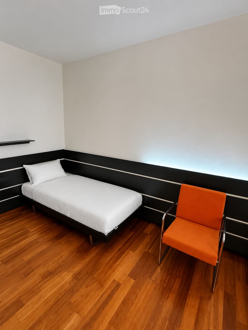Minimalist bedroom with a single bed, white bedding, and an orange chair, featuring wooden flooring and white walls.