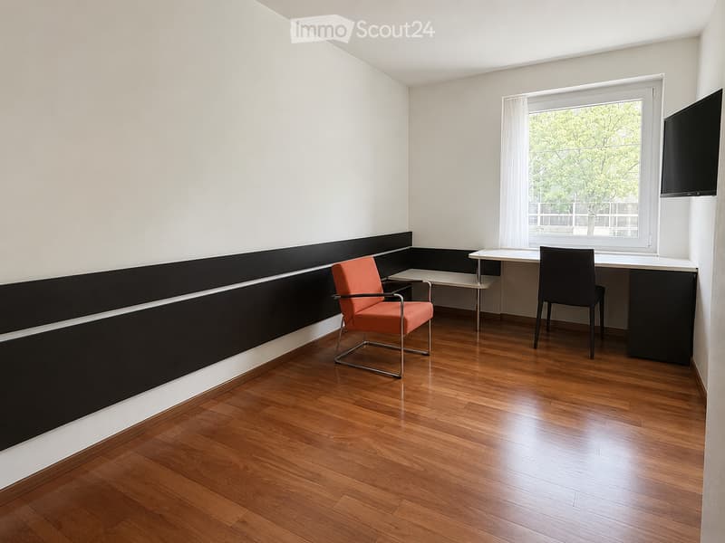 A modern, minimalist room with wooden flooring, white walls, and a large window with white curtains. Features include a black and white accent wall, a red chair, a small table, and a mounted screen.