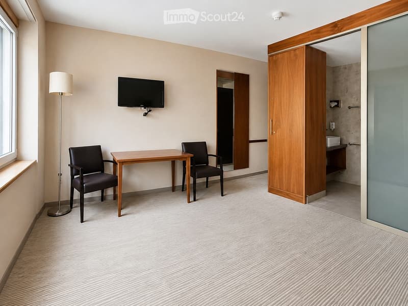 A furnished room with a desk, two chairs, a floor lamp, and a TV mounted on the wall. A wooden wardrobe and a glass-enclosed bathroom are visible.