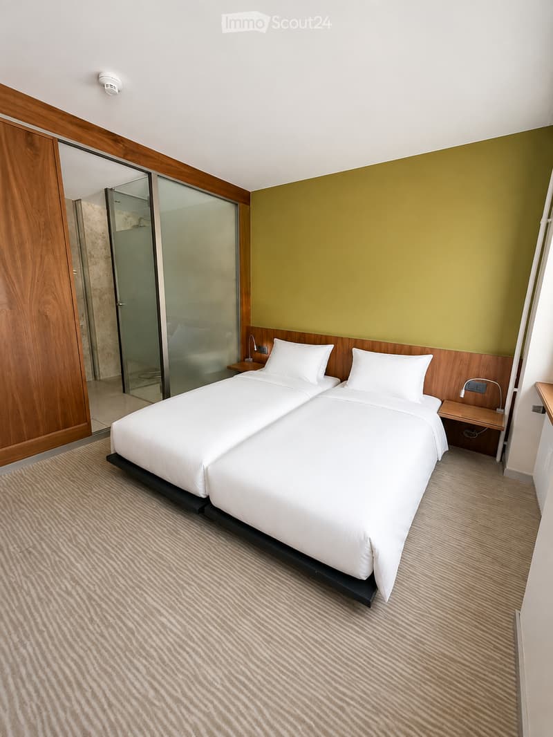 Modern bedroom with two single beds, white bedding, wooden nightstands, and a glass-enclosed shower area visible through an open doorway.