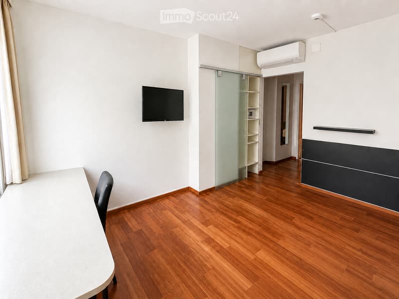 A modern, minimalist room with wooden flooring, a white table, a single chair, and a wall-mounted TV. Features a sliding glass door leading to another room.
