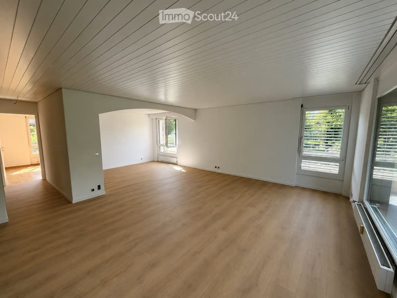 Empty room with wooden flooring, white walls, and large windows allowing natural light. The room features a slanted white ceiling and an open doorway leading to another space.
