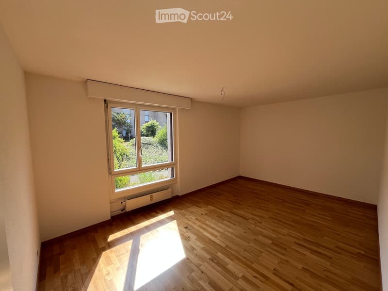 Empty room with wooden flooring, large window with blinds, radiator below window, and natural light streaming in.