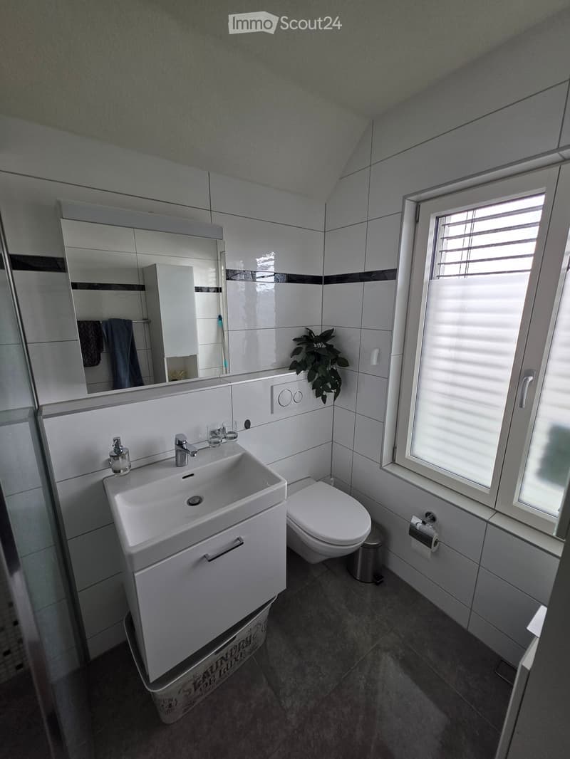 Modern bathroom with white tiles, a sink with a faucet, a toilet, a large mirror, and a small window with blinds.