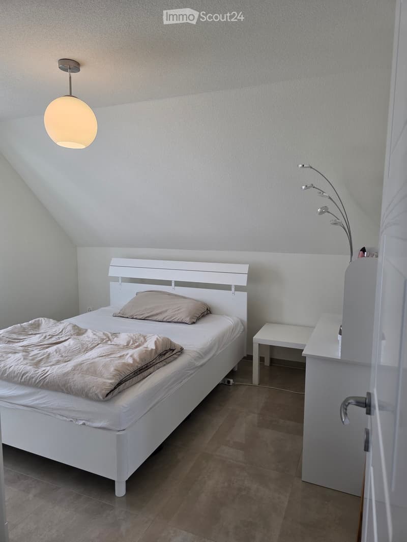 Modern bedroom with a white bed, beige bedding, a nightstand, and a floor lamp. The room features light-colored walls and a tiled floor.