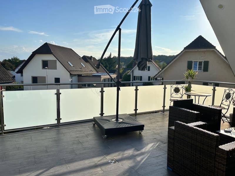 Balcony with wooden floor, glass railing, outdoor furniture including a table, chairs, and an umbrella.