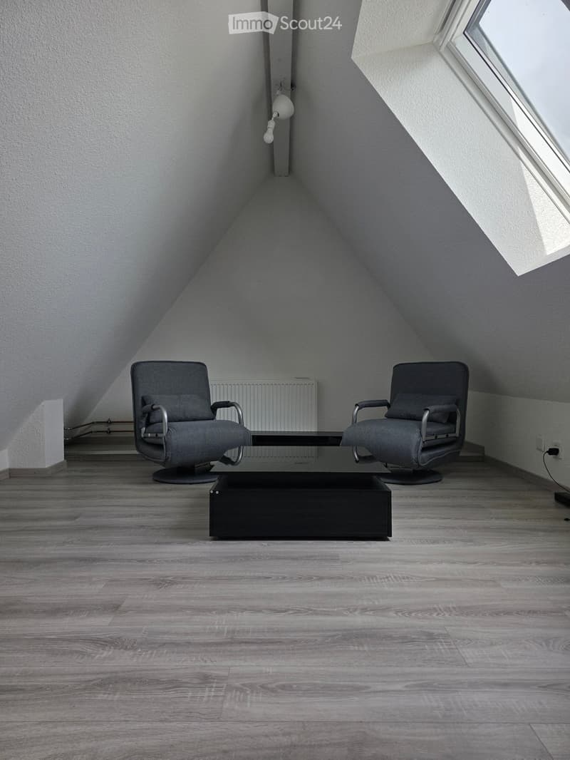 Modern attic room with wooden flooring, two grey armchairs, and a black coffee table.