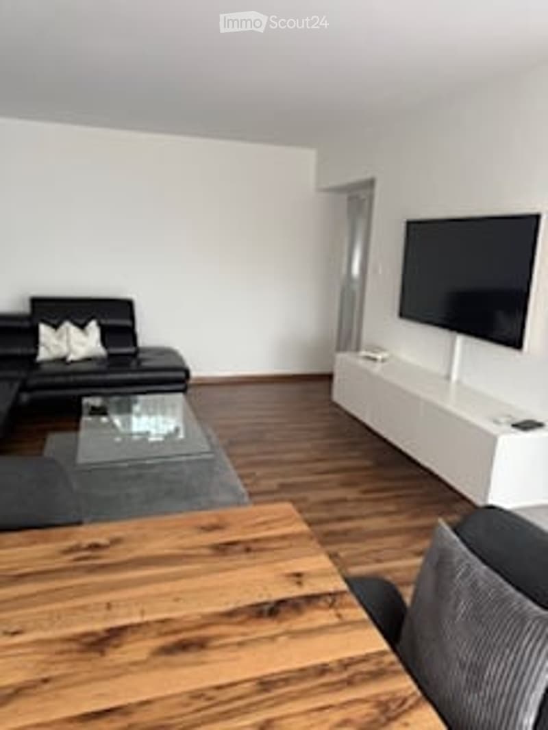 Modern living room with light wooden flooring, white walls, a black leather sofa, a glass coffee table, a TV on a white stand, and an ottoman.