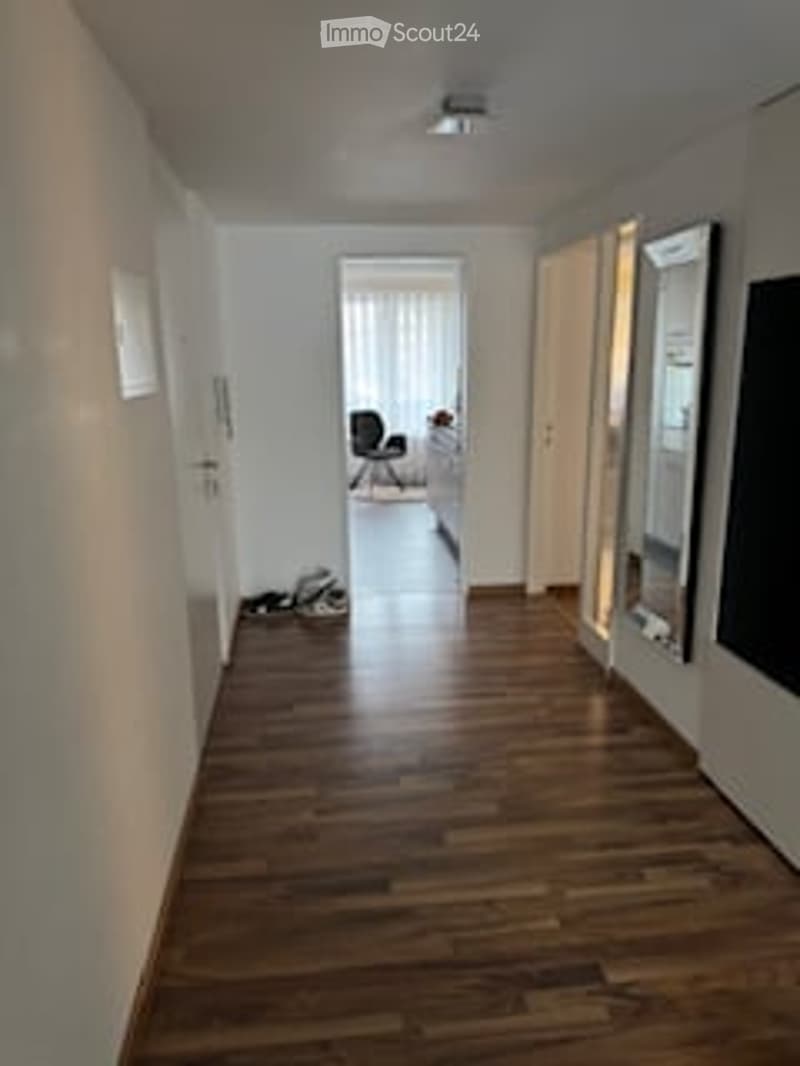 Empty hallway with wooden flooring, white walls, and a small seating area at the end.