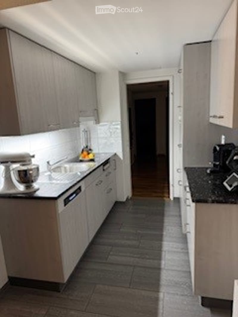 Modern kitchen with grey cabinets, black countertops, and a central island. Appliances include a sink, dishwasher, blender, and fridge. The floor is dark tiled.