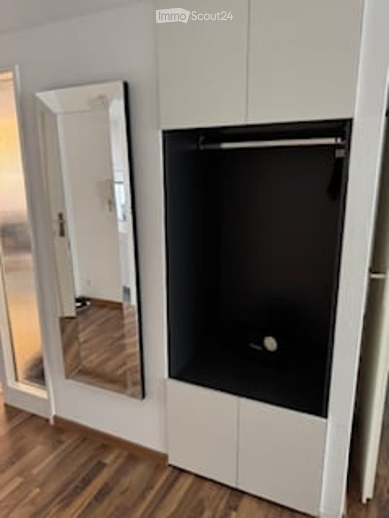 Modern room with a large mirror on the left and a flat-screen TV mounted on a white cabinet to the right. The floor is wooden, and the walls are white.