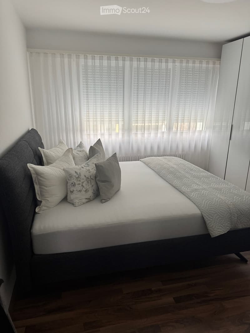 A modern bedroom with a neatly made bed, multiple pillows, a quilt, and a wardrobe. The room features wooden flooring and large windows with white curtains.