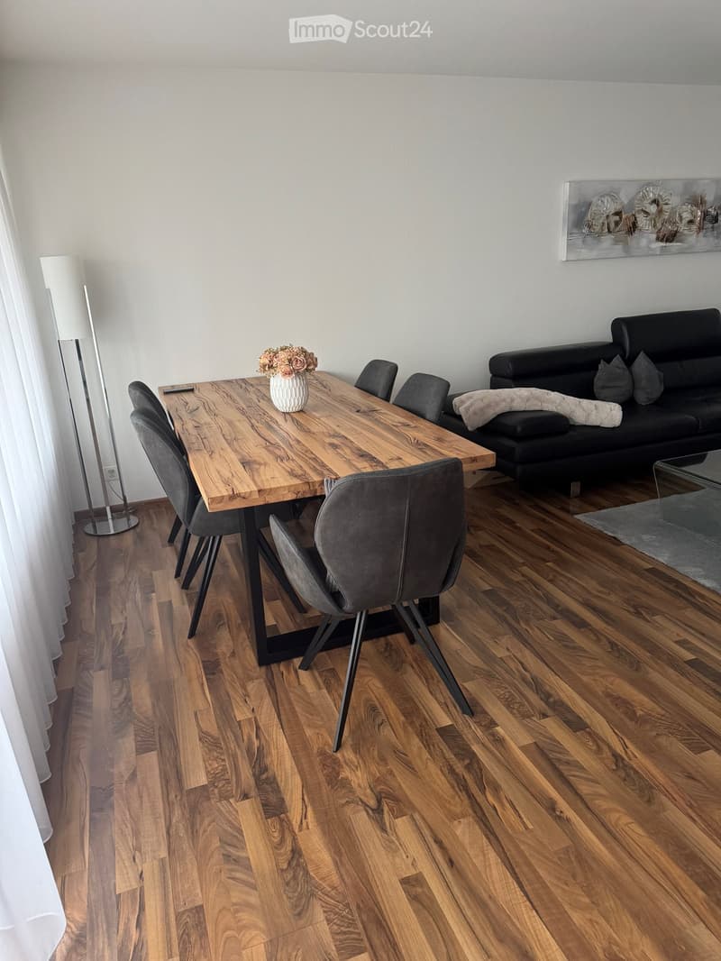 Modern living and dining area with wooden flooring, a black sofa, a wooden dining table with four chairs, a vase with flowers, and a floor lamp.