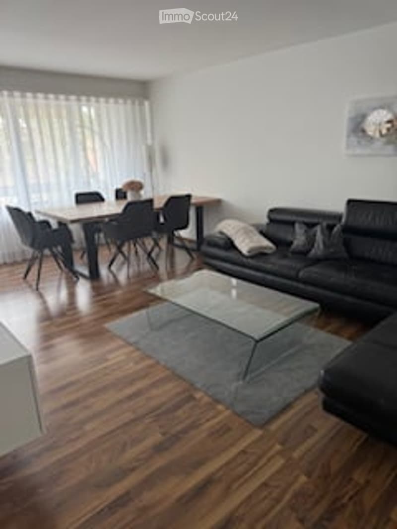 Bright, modern living and dining area with hardwood flooring, large windows with sheer curtains, a black sectional sofa, a glass coffee table, a dining table with six chairs, a floor lamp, and a decorative picture frame on the wall.