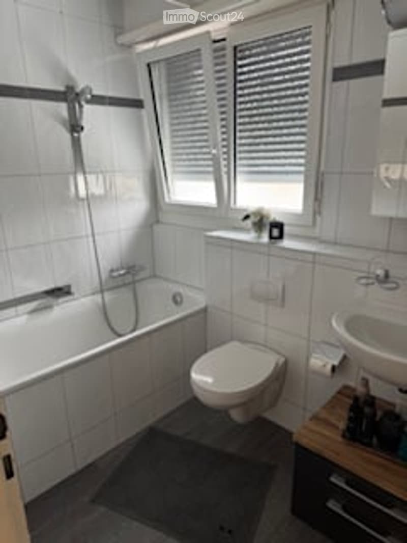 Modern bathroom with white tiles, a bathtub, toilet, sink, and a large window with blinds. Includes a mirror and a shower head.