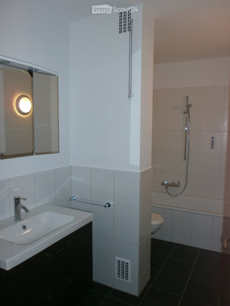 Modern bathroom with white tiles, a sink with a chrome faucet, a large mirror with a built-in light, a bathtub with a showerhead, and a toilet.