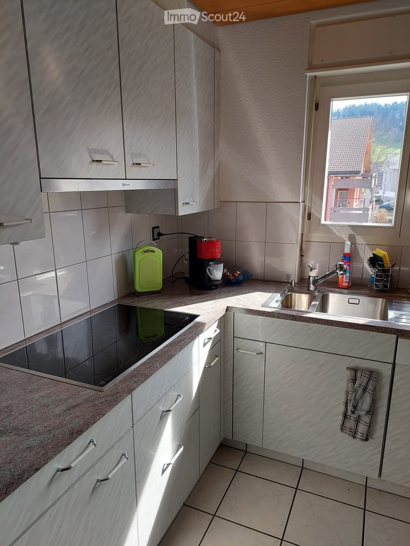 Modern kitchen with light-colored cabinets, stainless steel sink, and electric stovetop. Includes a coffee maker, cutting board, and towel.
