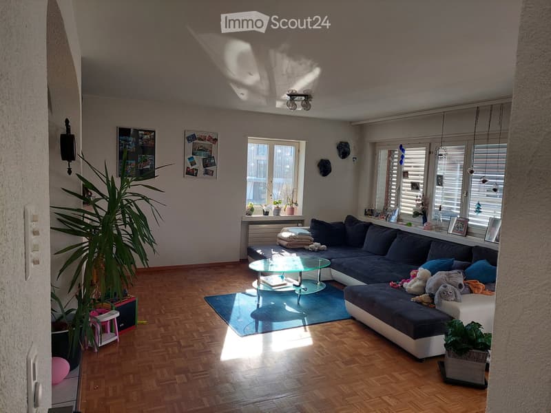 Bright living room with wooden flooring, large window, sectional sofa, glass coffee table, and decorative plants.