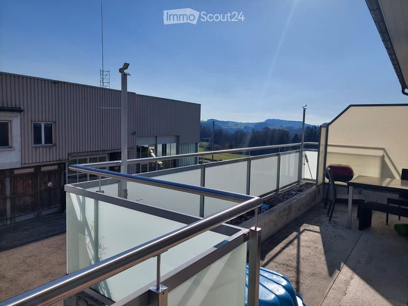 Balcony with metal railing, glass panels, small seating area with table, chairs, and grill, overlooking a landscape with buildings and hills.