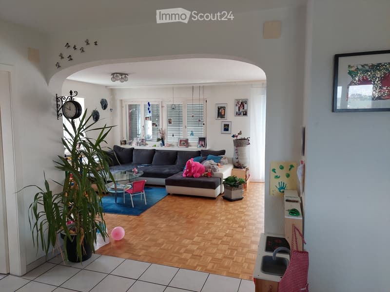 Bright living room with wooden flooring, large sectional sofa, glass coffee table, colorful chair, and decorative plants.