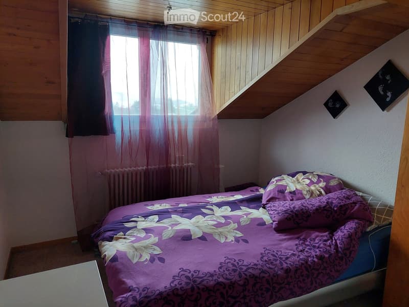 Cozy bedroom with a purple bedspread, floral design, and matching pillows. The room features wooden walls and a slanted ceiling, with a window covered by sheer curtains allowing natural light to enter.