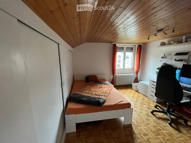 Cozy bedroom with a single bed, desk, chair, and shelves. The room features wooden flooring and a slanted wooden ceiling.