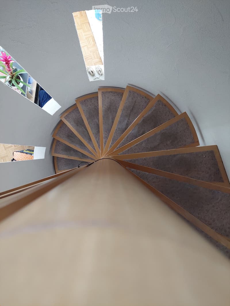 Spiral staircase with wooden handrails and carpeted steps, viewed from above, with grey walls and partial views of adjacent rooms.